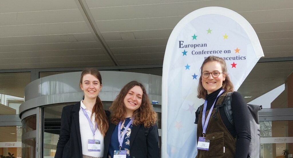 Three young women posing in front of the entrance to the European Conference on Pharmaceutics.