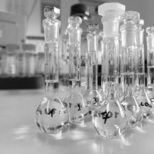 A black and white photo of a laboratory setting showing an array of glass volumetric flasks and graduated cylinders with stoppers, marked with handwritten labels. The lab equipment is arranged on a benchtop, reflecting the precision and cleanliness typical of a scientific research environment.