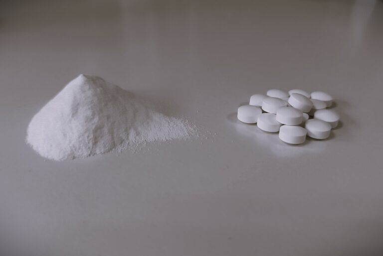 A monochromatic image showcasing a neat pile of white powder on the left and a group of round white tablets on the right, arranged on a smooth surface. The composition conveys a stark contrast between the raw form of a substance and its processed tablet form.