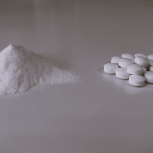 A monochromatic image showcasing a neat pile of white powder on the left and a group of round white tablets on the right, arranged on a smooth surface. The composition conveys a stark contrast between the raw form of a substance and its processed tablet form.
