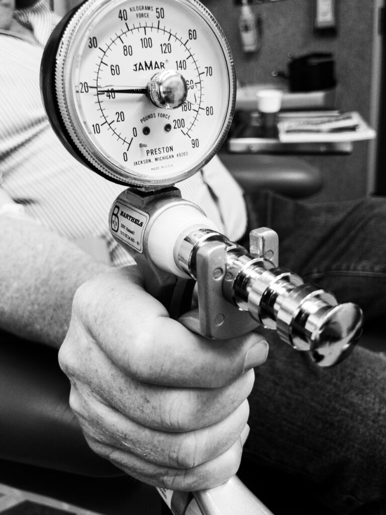 A detailed black and white photograph captures an elderly individual's hand engaging a Jamar hand dynamometer, a clinical tool designed to evaluate hand grip strength. The gauge's needle indicates a measurement, with dual scales in both kilograms and pounds force. The dynamometer's metallic components and textured grip handle are prominently displayed as the person's hand exerts pressure, showcasing a moment of strength assessment in a clinical or rehabilitative setting.