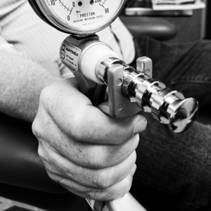 A detailed black and white photograph captures an elderly individual's hand engaging a Jamar hand dynamometer, a clinical tool designed to evaluate hand grip strength. The gauge's needle indicates a measurement, with dual scales in both kilograms and pounds force. The dynamometer's metallic components and textured grip handle are prominently displayed as the person's hand exerts pressure, showcasing a moment of strength assessment in a clinical or rehabilitative setting.