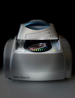 A laboratory qPCR machine, the Rotor-Gene Q, is shown with a sleek design featuring a metallic silver exterior and a prominent blue-lit lid. The machine is situated against a gradient grey background, highlighting its modern and sophisticated aesthetic.