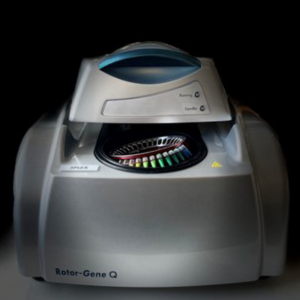 A laboratory qPCR machine, the Rotor-Gene Q, is shown with a sleek design featuring a metallic silver exterior and a prominent blue-lit lid. The machine is situated against a gradient grey background, highlighting its modern and sophisticated aesthetic.